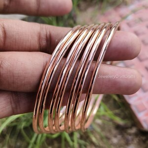 May include: A set of copper-coloured bangles, held in a hand. The bangles are thin, round, and have a polished finish. The image is taken in natural light, highlighting the metallic sheen of the jewellery. The bangles are stacked together, showcasing their design.