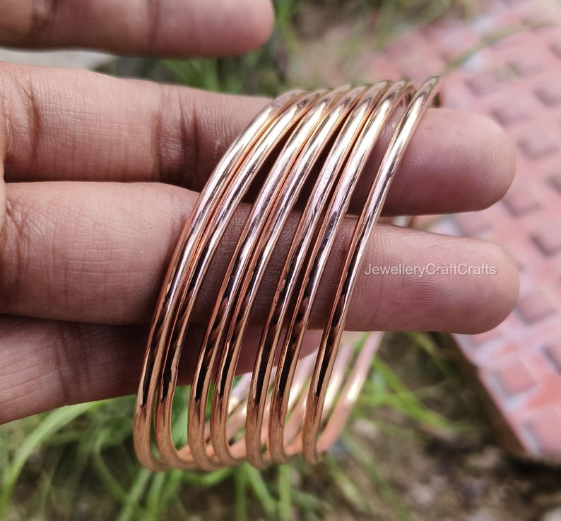 May include: A set of copper-coloured bangles, held in a hand. The bangles are thin, round, and have a polished finish. The image is taken in natural light, highlighting the metallic sheen of the jewellery. The bangles are stacked together, showcasing their design.