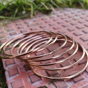 May include: A collection of thin, rose gold bangles. The bangles are circular and have a smooth, polished finish. The jewellery is resting on a reddish-brown, textured surface with green grass in the background. The bangles are stacked together.