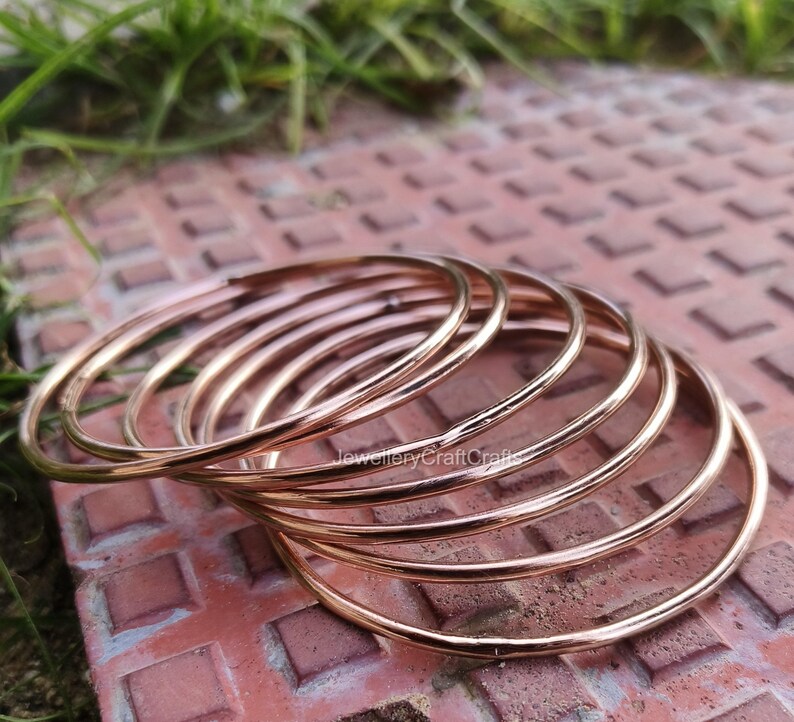 May include: A collection of thin, rose gold bangles. The bangles are circular and have a smooth, polished finish. The jewellery is resting on a reddish-brown, textured surface with green grass in the background. The bangles are stacked together.