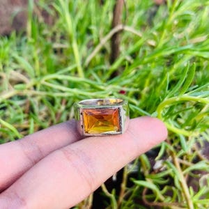 Könnte beinhalten: Ein silberfarbener Ring mit einem großen, rechteckigen, facettierten bernsteinfarbenen Edelstein, der in eine breite, polierte Metallschiene gefasst ist. Der Ring hat eine massive, solide Konstruktion mit einem klaren, leuchtend orange-gelben Stein.