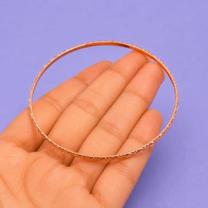Könnte beinhalten: Ein dünner, kreisförmiger Armreif aus Roségold mit einem feinen, sich wiederholenden eingravierten Muster auf der gesamten Außenfläche. Das Metall hat eine polierte, reflektierende Oberfläche, die das Licht einfängt und eine subtile Textur erzeugt.