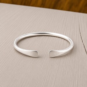 Könnte beinhalten: Ein silberfarbenes Armband in offener Form mit einer glatten, polierten Oberfläche. Der kreisförmige Reifen weist vorne eine Öffnung auf, mit zwei abgerundeten, flachen Enden, die leicht zulaufen. Das Metall hat einen gleichmäßigen Glanz.