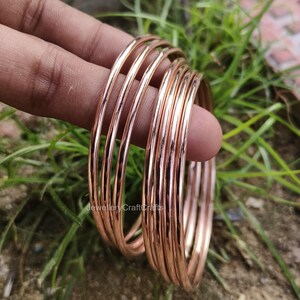 May include: A set of thin, rose gold bangles. The bangles are stacked together, reflecting light. The image is set against a backdrop of green grass and foliage. The bangles are a simple, elegant design.