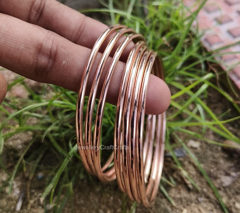 May include: A set of thin, rose gold bangles. The bangles are stacked together, reflecting light. The image is set against a backdrop of green grass and foliage. The bangles are a simple, elegant design.