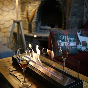 May include: A rectangular tabletop fireplace with a black base and clear glass panels, featuring a central metal burner with active flames. Two stemmed wine glasses containing a brown liquid are positioned on either side of the fireplace.