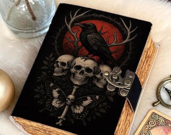 May include: A black journal with a gothic design featuring a raven, skulls, and a butterfly. The cover has a decorative metal clasp and aged-look edges. The journal is on a white, fluffy surface.