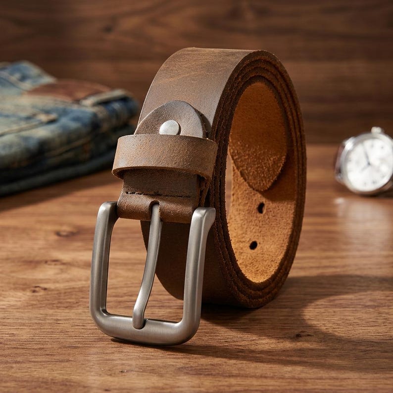 Puede incluir: Un cintur&oacute;n de cuero marr&oacute;n enrollado con una hebilla plateada. El cintur&oacute;n est&aacute; sobre una superficie de madera, con un par de vaqueros y un reloj en el fondo. El cintur&oacute;n tiene un aspecto cl&aacute;sico y robusto. El ancho del cintur&oacute;n es de unos 3,8 cm.