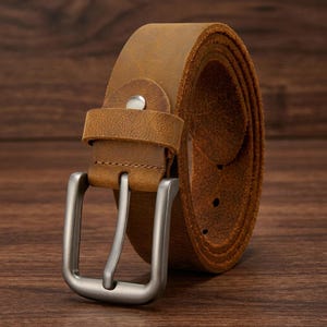 Puede incluir: Un cintur&oacute;n de cuero marr&oacute;n enrollado con una hebilla plateada. El cintur&oacute;n presenta un dise&ntilde;o cl&aacute;sico con una hebilla rectangular y una presilla para asegurar el extremo. El cuero tiene un aspecto envejecido, lo que aumenta su atractivo vintage. El cintur&oacute;n mide entre 70 cm y 100 cm.