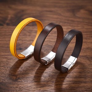 May include: Three leather bracelets in yellow, brown, and black, each with a silver magnetic clasp. The bracelets are displayed on a wooden surface, showcasing their simple, modern design. The leather appears smooth and high-quality.