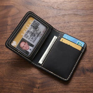 Op de afbeelding: Zwart leren bi-fold portemonnee met witte stiksels. De portemonnee is open en onthult een doorzichtig ID-venster met een foto en tekst, en sleuven voor kaarten en contant geld. De ID luidt "S.H.I.E.L.D."