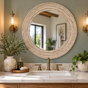 May include: A round wall mirror with a textured, light-colored woven frame hangs above a white marble bathroom vanity. The vanity features a brass faucet, a ceramic vase with greenery, a soap dispenser, and a potted plant.