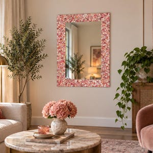 May include: A rectangular wall mirror featuring a wide frame covered in a mosaic of small, overlapping pink and white shell-like pieces. The reflective glass surface is clear and smooth, displaying a reflection of the surrounding room interior.