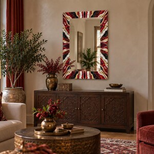 May include: A rectangular wall mirror featuring a decorative frame with a radial pattern of red, black, white, and gold-toned segments. The mirror is mounted on a wall above a dark wooden sideboard, reflecting the interior space of a room.