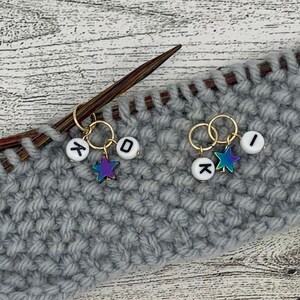 Op de afbeelding: Twee gouden brei-steekmarkeerders met witte letterparels en iriserende sterbedels. De kralen spellen "K" en "O" op de ene markeerder en "I" en "X" op de andere. De markeerders zijn bevestigd aan een grijze gebreide stof.