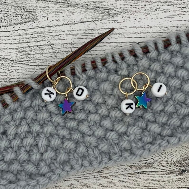 Op de afbeelding: Twee gouden brei-steekmarkeerders met witte letterparels en iriserende sterbedels. De kralen spellen "K" en "O" op de ene markeerder en "I" en "X" op de andere. De markeerders zijn bevestigd aan een grijze gebreide stof.