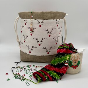 May include: A white drawstring bag with a reindeer pattern, a green and red striped knitted sock, and a small white bag with a reindeer design. The bag is filled with stitch markers and a charm.
