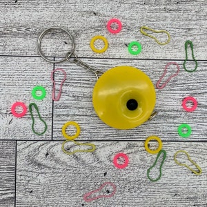 May include: A yellow circular tape measure with a black center, attached to a keychain. Scattered around are colorful stitch markers and ring markers in pink, green, and yellow, on a wood-grain surface.