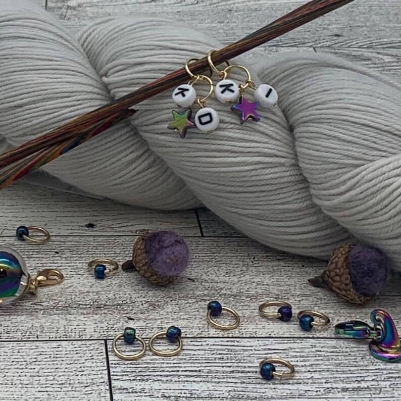 Op de afbeelding: Een close-up van een bol wit garen met gouden brei-markeringen met letter kralen en ster kralen. De brei-markeringen zitten op een houten breinaald. Er zijn ook verschillende gouden en blauwe brei-markeringen op een houten oppervlak.