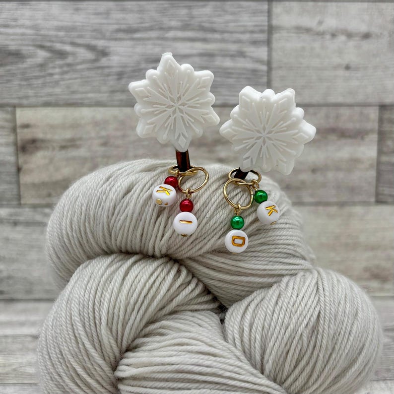 May include: Two white snowflake stitch markers with gold rings and beads. The beads are white with gold letters, "K" and "D", and have red and green accents.