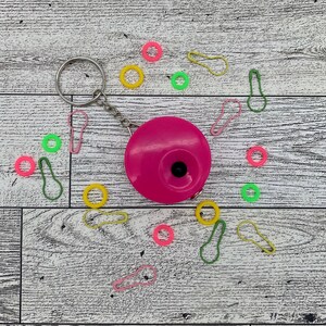May include: A bright pink circular tape measure with a silver keychain, surrounded by colorful stitch markers in pink, yellow, and green. The items are arranged on a white and gray wooden surface.