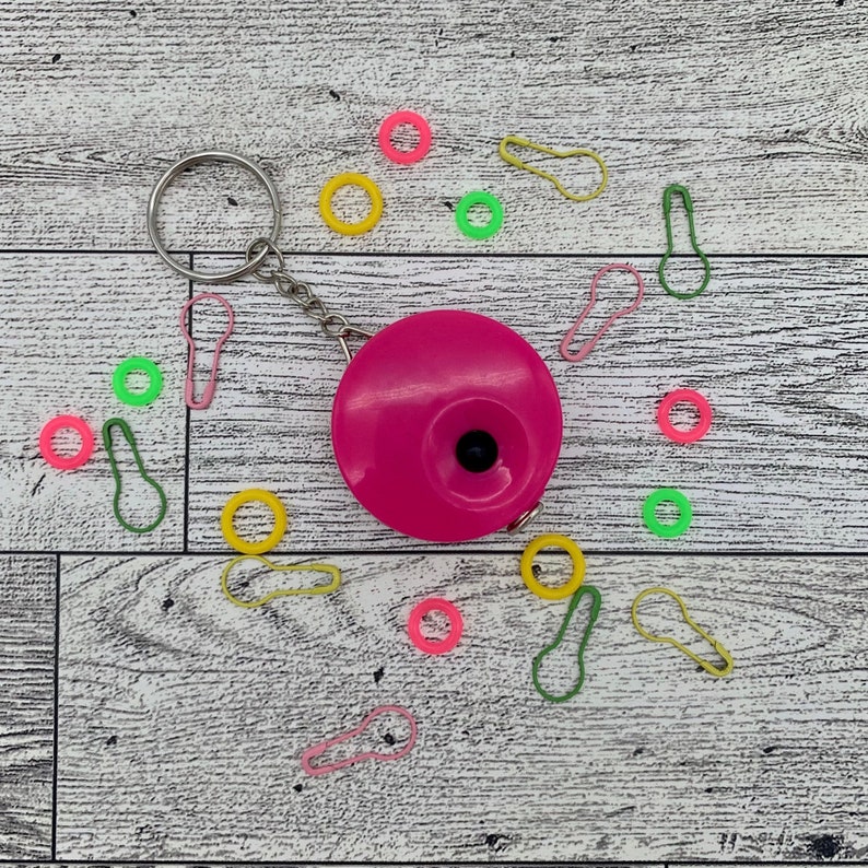 May include: A bright pink circular tape measure with a silver keychain, surrounded by colorful stitch markers in pink, yellow, and green. The items are arranged on a white and gray wooden surface.