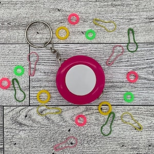 May include: A hot pink retractable tape measure with a white center, attached to a silver keyring. Scattered around are colorful stitch markers in pink, yellow, and green, and round stitch markers in pink, yellow, and green.