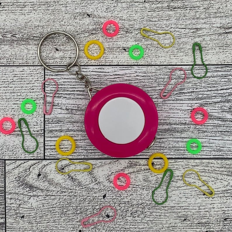 May include: A hot pink retractable tape measure with a white center, attached to a silver keyring. Scattered around are colorful stitch markers in pink, yellow, and green, and round stitch markers in pink, yellow, and green.