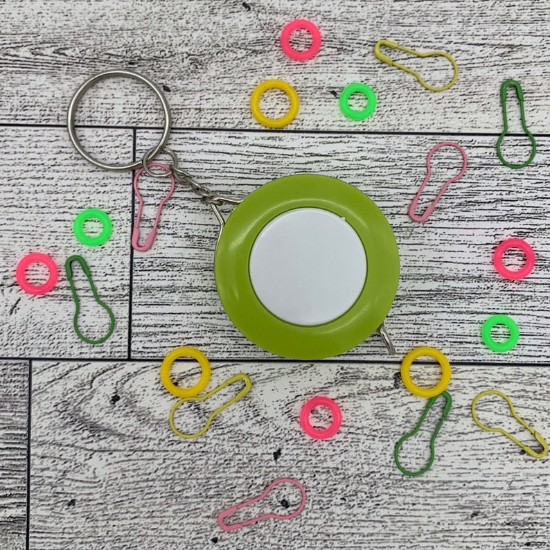 May include: A green and white round measuring tape with a silver keychain, surrounded by colorful stitch markers in pink, yellow, and green. The stitch markers are in the shape of safety pins and rings.