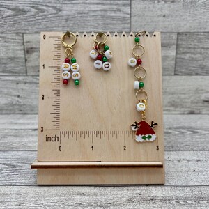 May include: Three Christmas themed stitch markers with gold rings and beads. The markers are on a wooden ruler with inch markings. The first marker has red and green beads with the letters "RSS" on the beads. The second marker has red and green beads with the letters "KID" on the beads. The third marker has red and green beads with a small Santa hat charm.