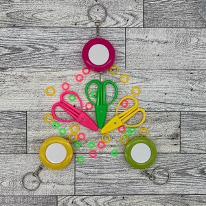 May include: A collection of colorful craft supplies, including pink, green, and yellow scissors, along with matching keychains and small, colorful stitch markers. The items are arranged on a light wood-grain surface.