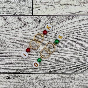 May include: Two gold chain necklaces with white beads that have gold letters on them. The beads are red and green. The necklaces are on a light wood background.