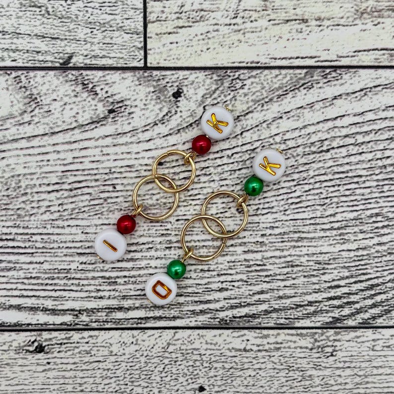 May include: Two gold chain necklaces with white beads that have gold letters on them. The beads are red and green. The necklaces are on a light wood background.