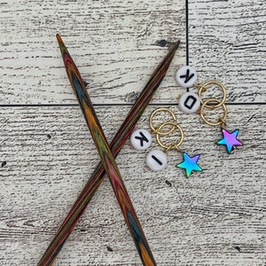 May include: Two wooden knitting needles crossed over each other. Three white round stitch markers with black letters "K", "I", and "O" are on a white wooden surface. Two gold ring stitch markers with iridescent blue star charms are also on the surface.