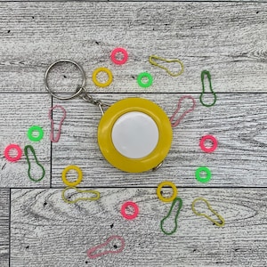 May include: A yellow measuring tape with a white center and a silver key ring. Scattered around are colorful stitch markers in pink, green, and yellow. The items are arranged on a light wood-grain surface.