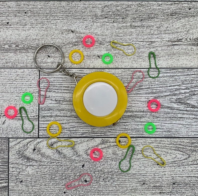 May include: A yellow measuring tape with a white center and a silver key ring. Scattered around are colorful stitch markers in pink, green, and yellow. The items are arranged on a light wood-grain surface.