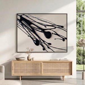 May include: Abstract art piece featuring black paint splatters on a neutral canvas, framed in black. The artwork hangs above a light wood cabinet with woven doors, creating a modern interior design aesthetic.
