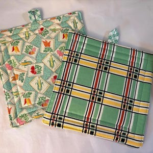 May include: Two square quilted pot holders with fabric loops. One features a light green background with a repeating pattern of vegetables in diamond frames. The other displays a green, yellow, black, and red plaid pattern with quilted stitching.