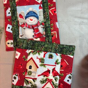 May include: Two square quilted pot holders featuring a snowman in a blue hat and red scarf, red cardinals, and birdhouses. The fabric has a red background with green pine branch borders and a small fabric loop at the top for hanging.