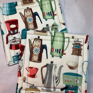 May include: Two square fabric pot holders featuring a repeating pattern of various coffee-making equipment. The illustrations include brown coffee pots with white floral designs, green kettles, red coffee makers, and brown manual coffee grinders.