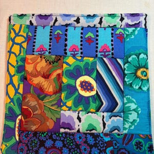 May include: A square fabric potholder featuring a patchwork design of various colorful floral and geometric patterns in shades of blue, green, yellow, purple, and red. The quilted piece is constructed from multiple rectangular and square fabric segments.