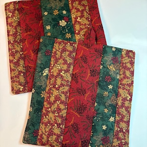 May include: Two square quilted pot holders featuring a vertical striped pattern. The fabric displays alternating sections of red with pinecone motifs, dark green with floral patterns, and tan with dense botanical prints. Each has a small fabric hanging loop.