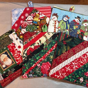 May include: A collection of five quilted fabric pot holders featuring various holiday themes. Designs include a Santa Claus portrait, a group of four snowmen, and several patchwork patterns in red, green, and white with holly and floral prints.