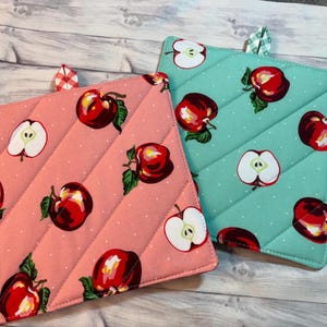 May include: Two quilted pot holders with apple patterns. One is pink with red apples and white polka dots, the other is teal with red apples and white polka dots. Both have a loop for hanging.