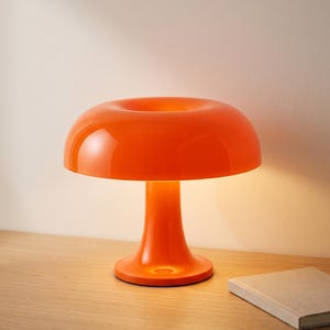May include: A vibrant orange mushroom-shaped table lamp. The lamp features a rounded shade with a central opening, a slender stem, and a circular base. The glossy finish reflects light, creating a warm glow. The lamp is on a wooden surface.