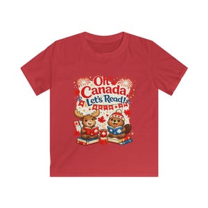 May include: A red short-sleeved t-shirt featuring a graphic of a cartoon moose and beaver reading books. The text reads Oh Canada, Let's Read! The design includes maple leaves, fireworks, a small Canadian flag, and bunting in red, white, and blue.