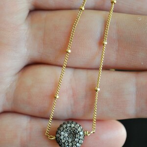 May include: A delicate gold-colored necklace featuring a round pendant encrusted with small, sparkling stones. The chain has a fine, textured design with small, evenly spaced gold beads. The pendant is the focal point, catching the light and adding a touch of elegance.