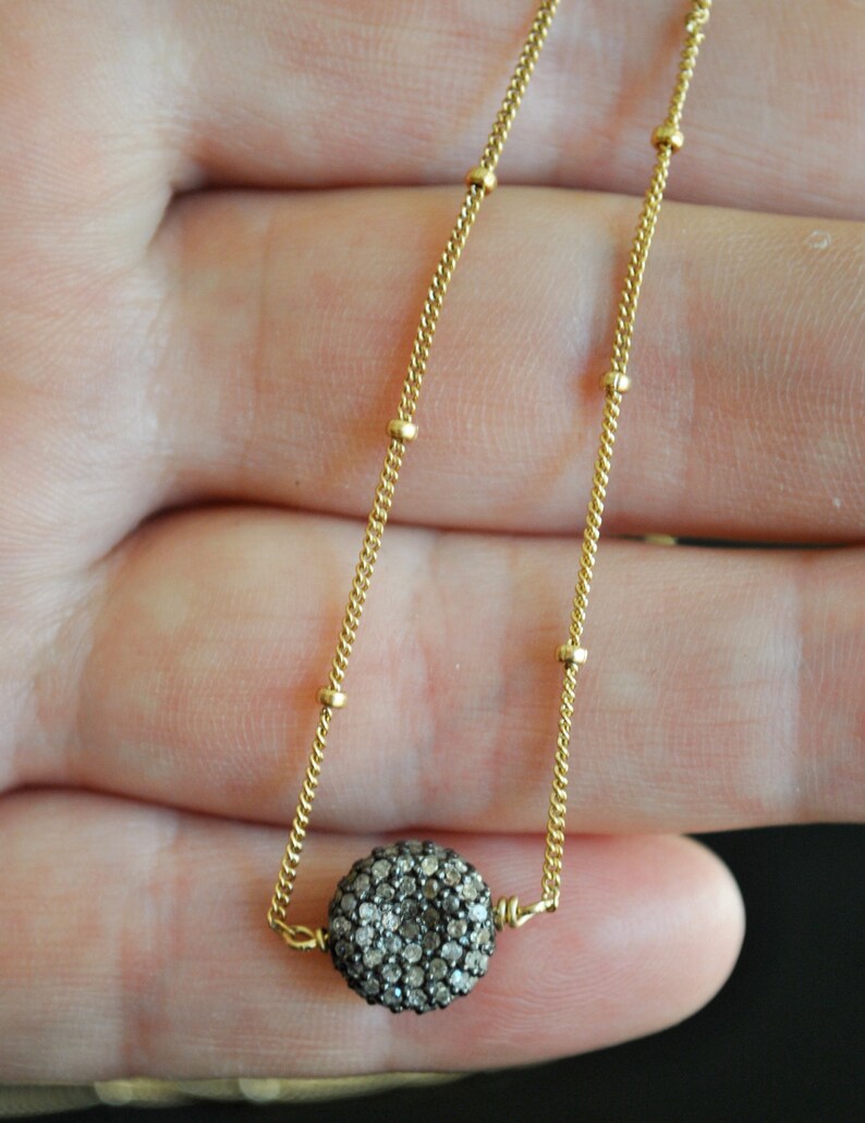May include: A delicate gold-colored necklace featuring a round pendant encrusted with small, sparkling stones. The chain has a fine, textured design with small, evenly spaced gold beads. The pendant is the focal point, catching the light and adding a touch of elegance.