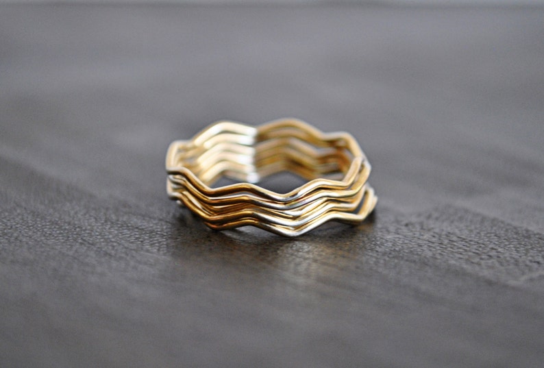 May include: A set of five gold and silver wavy rings stacked together. The rings are made of thin metal and have a simple, modern design.