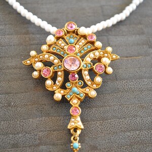 May include: A gold necklace with a pink and turquoise pendant featuring a detailed ornate design. The pendant is shaped like a teardrop and has a pearl-like finish.